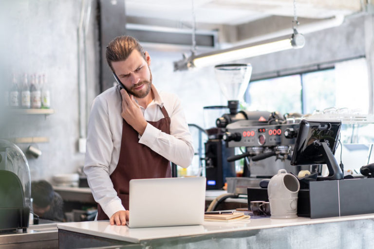 Male barista next to coffee machine and with computer in front of him frowning whilst on the phone