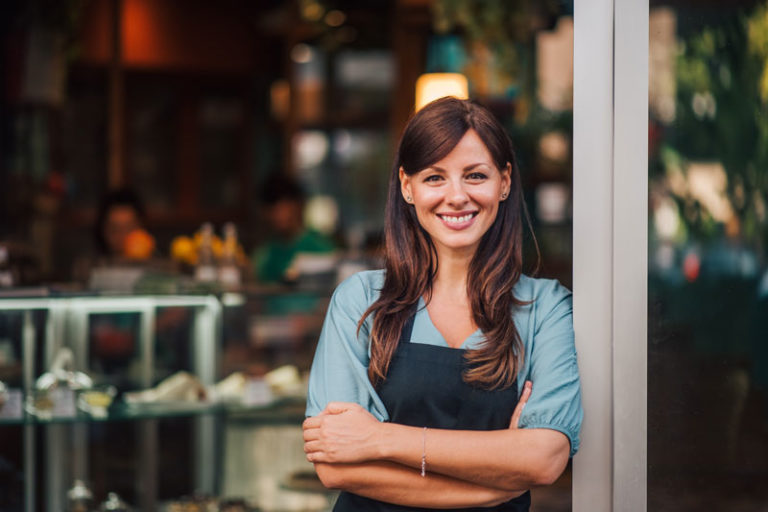 Mid-shot of standing female in apron smiling and folding her arms whilst leaning against the entry way to a store.